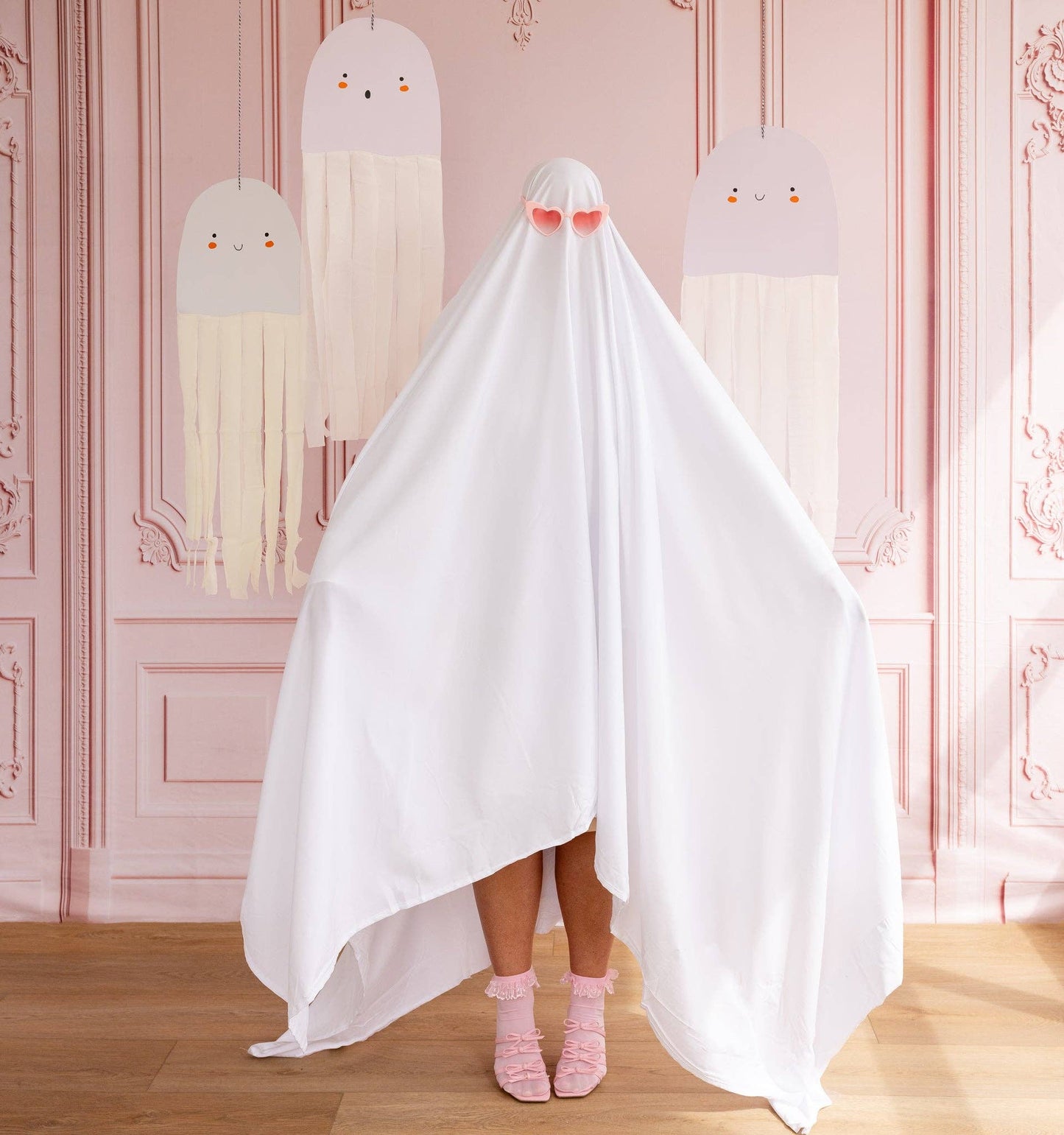 Person wearing a white ghost costume with pink socks in a room with decorative pink walls and hanging ghost decor.