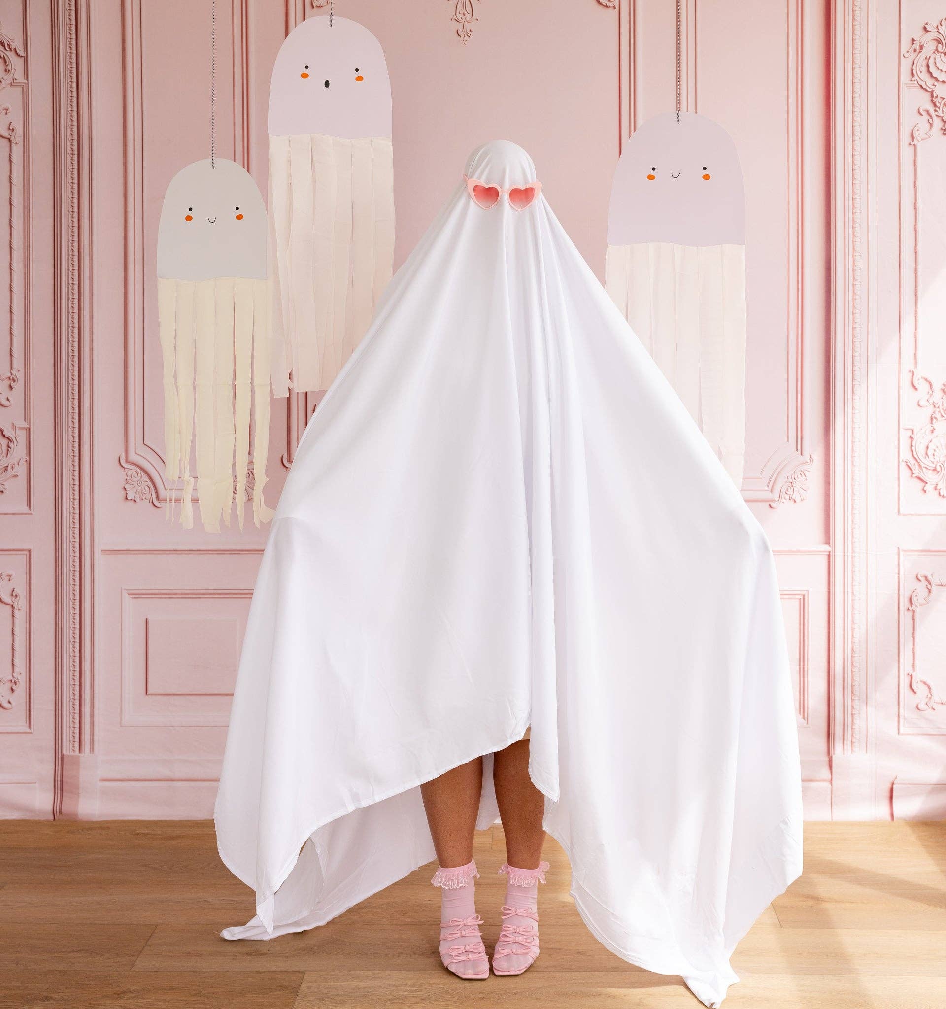 Person wearing a white ghost costume with pink socks in a room with decorative pink walls and hanging ghost decor.