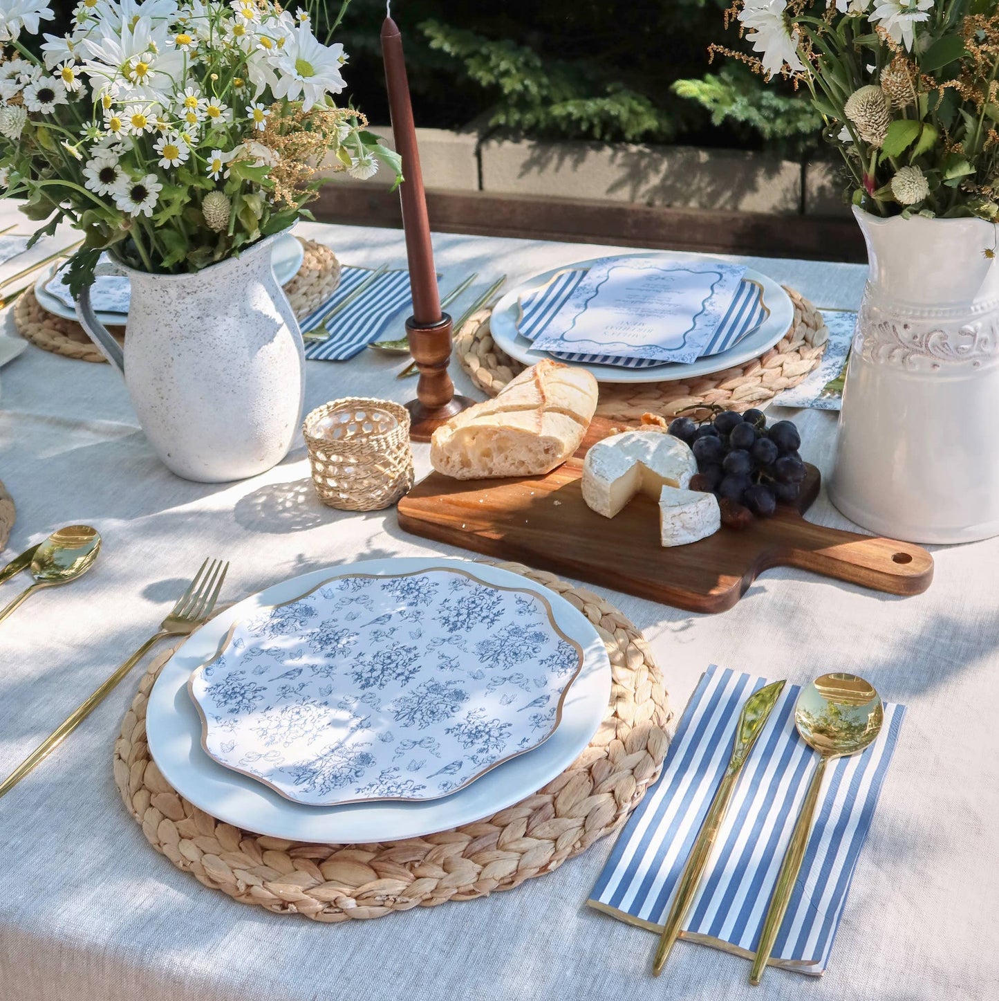 Outdoor table setting with plates, bread, cheese, and flowers on a white tablecloth.