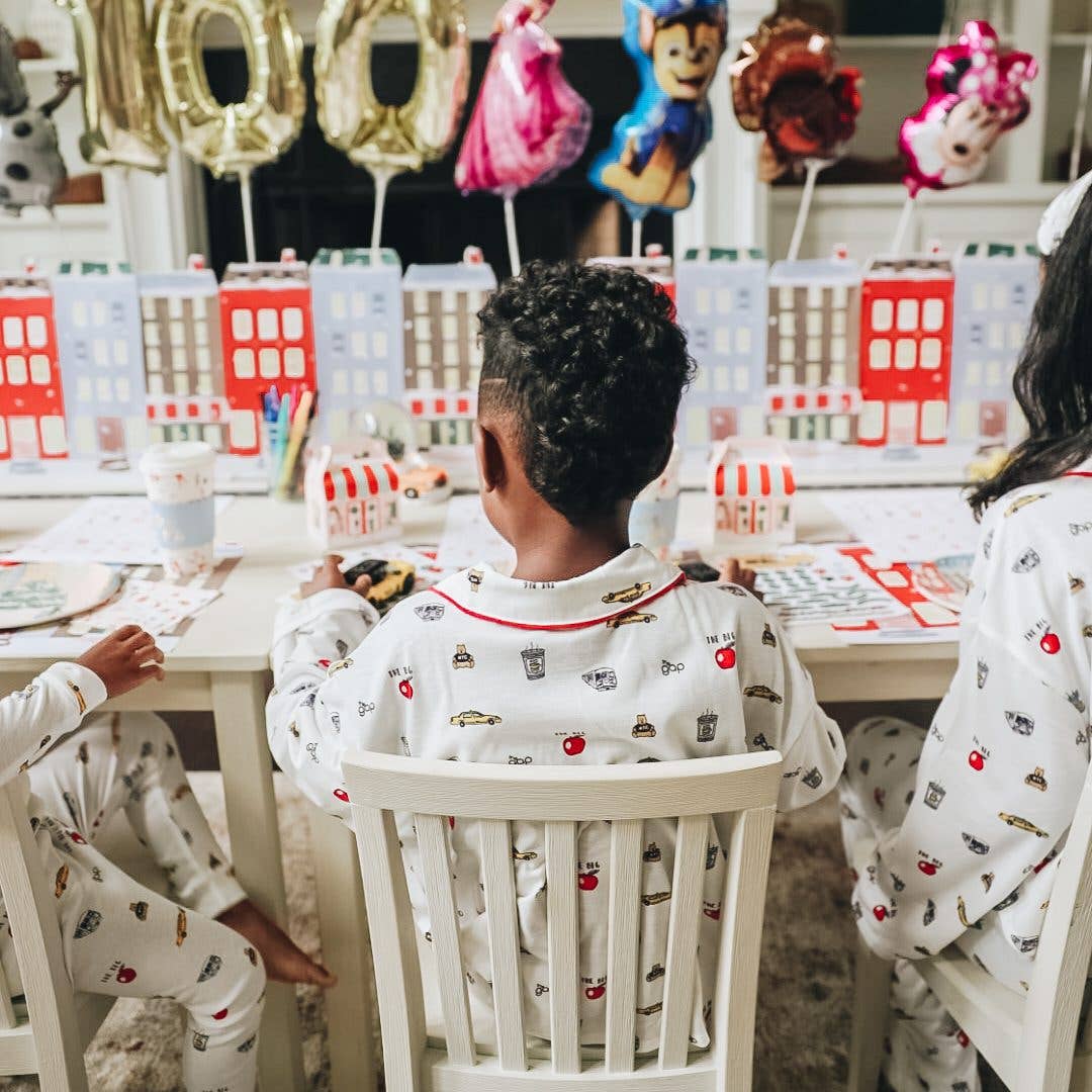 Three children sitting at a table from behind, wearing matching white Christmas pajamas with a colorful cartoon food and drink print. The room is decorated for a holiday party with house-shaped paper cutouts, colorful balloons, and party supplies.