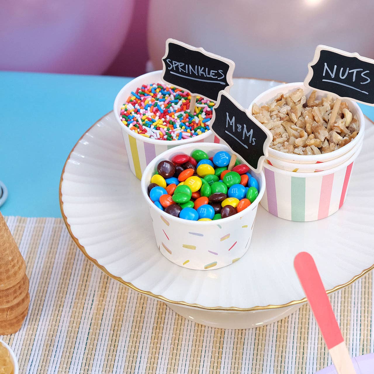 Wooden chalkboard toppers labeled 'SPRINKLES' and 'NUTS' placed in colorful paper bowls filled with sprinkles, M&Ms, and nut pieces, with a party atmosphere.