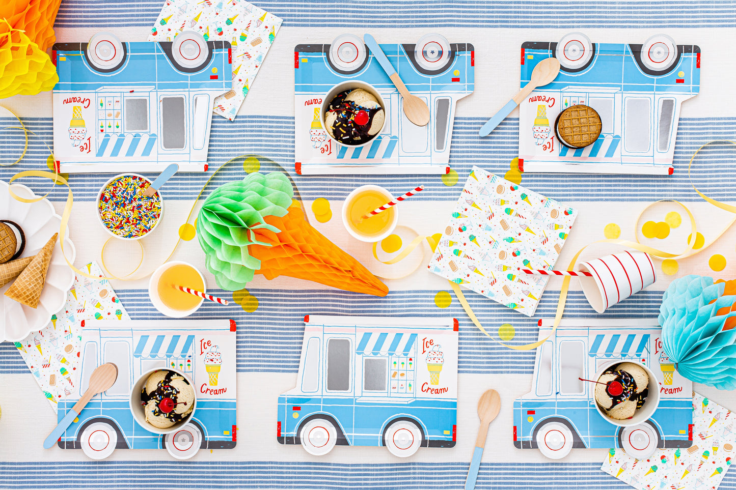 A flat-lay view of an ice cream truck themed party table setting, featuring printed paper napkins, cups, straws, and decorative paper fans in light blue, white, and multicolor patterns.