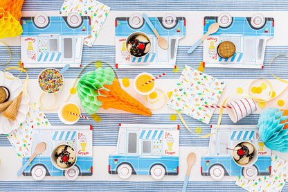A flat-lay view of an ice cream truck themed party table setting, featuring printed paper napkins, cups, straws, and decorative paper fans in light blue, white, and multicolor patterns.