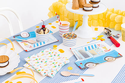 A styled ice cream party table setting with matching printed napkins, cups, straws, paper plates, and dessert props on a white tablecloth with blue stripes and yellow accents.
