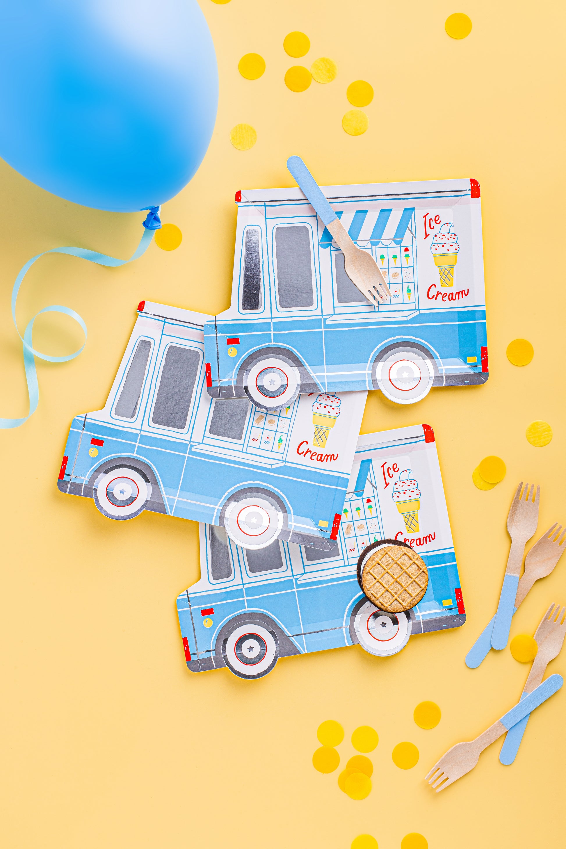 A flat-lay of light blue and white paper ice cream truck party decorations on a yellow background, including printed napkins, plastic forks, and a blue balloon with confetti.