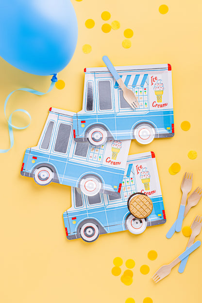 A flat-lay of light blue and white paper ice cream truck party decorations on a yellow background, including printed napkins, plastic forks, and a blue balloon with confetti.