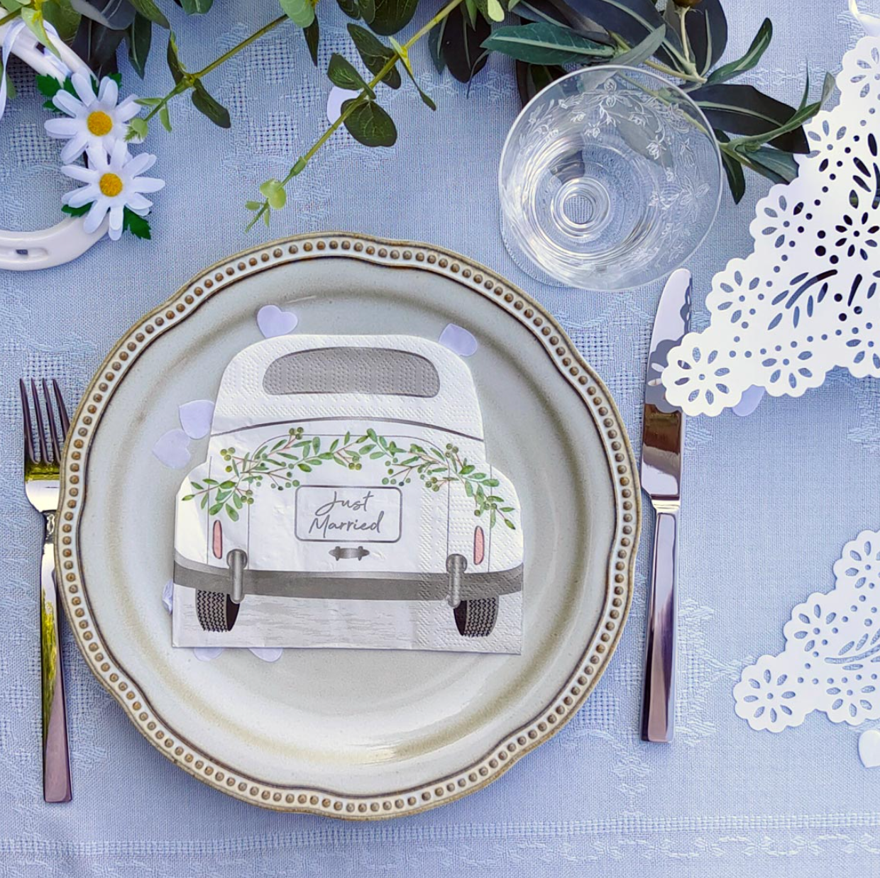 A styled place setting on a light blue tablecloth featuring a decorative ceramic plate with a vintage-style wedding napkin in the center. The napkin is printed with a cream-colored car adorned with green vines and the words 'Just Married.