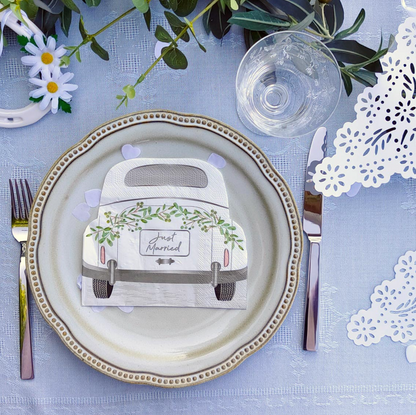 A styled place setting on a light blue tablecloth featuring a decorative ceramic plate with a vintage-style wedding napkin in the center. The napkin is printed with a cream-colored car adorned with green vines and the words 'Just Married.
