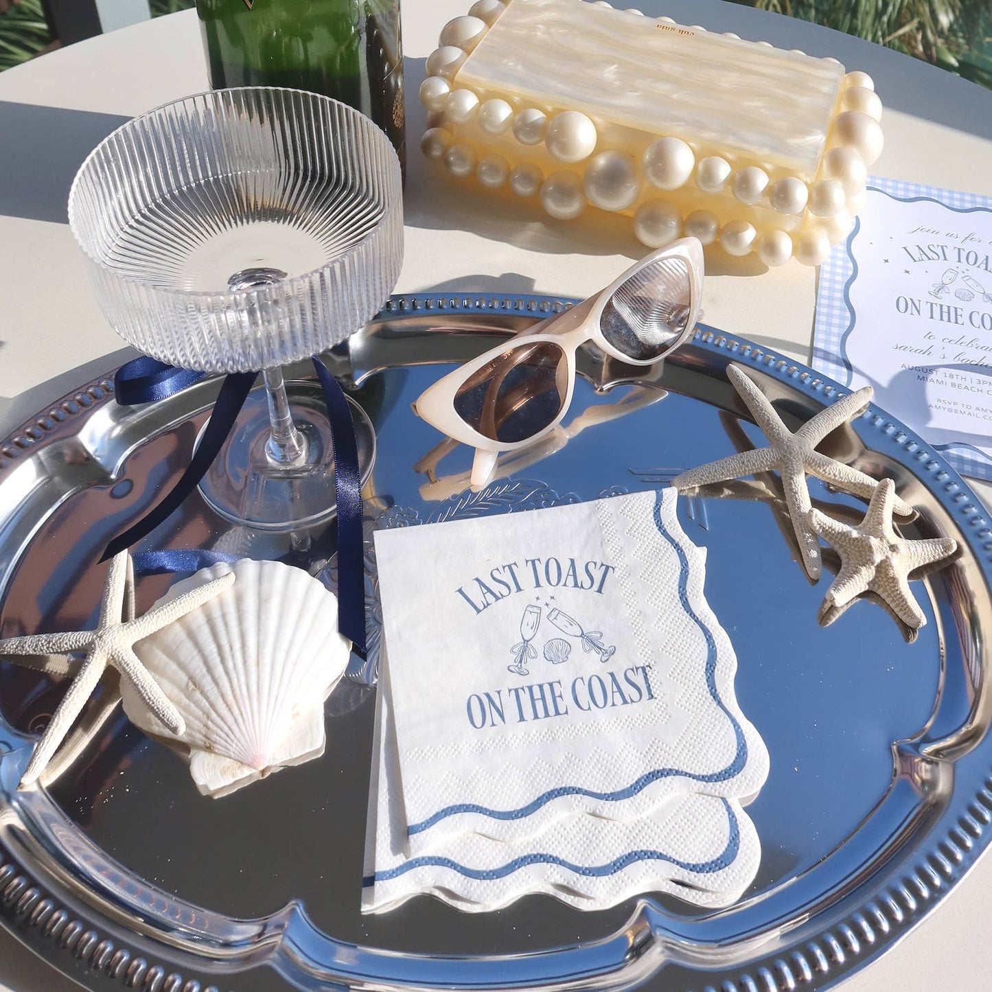 Decorative tray with 'Last Toast on the Coast' card, sunglasses, and shell decorations on a table.