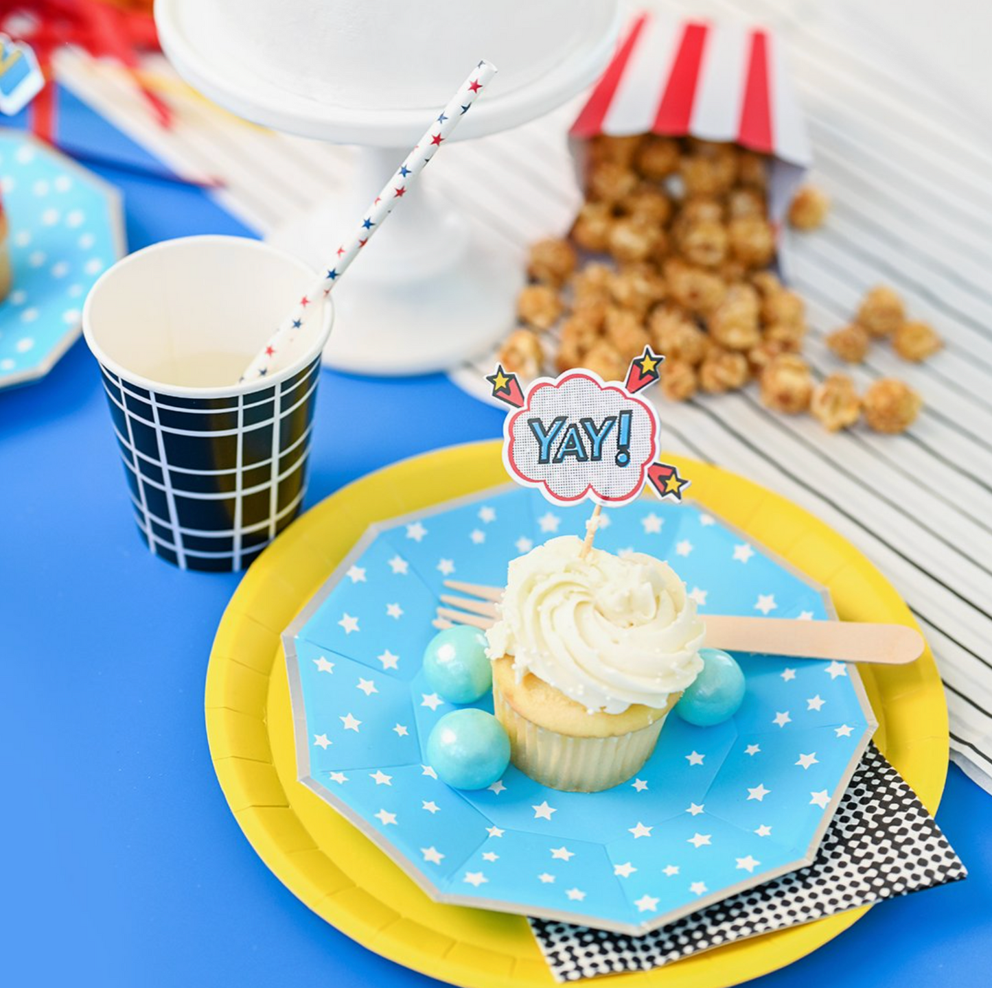 Cupcake on a blue plate with 'YAY!' topper, surrounded by party supplies on a blue tablecloth, including a paper straw with red and blue stars.