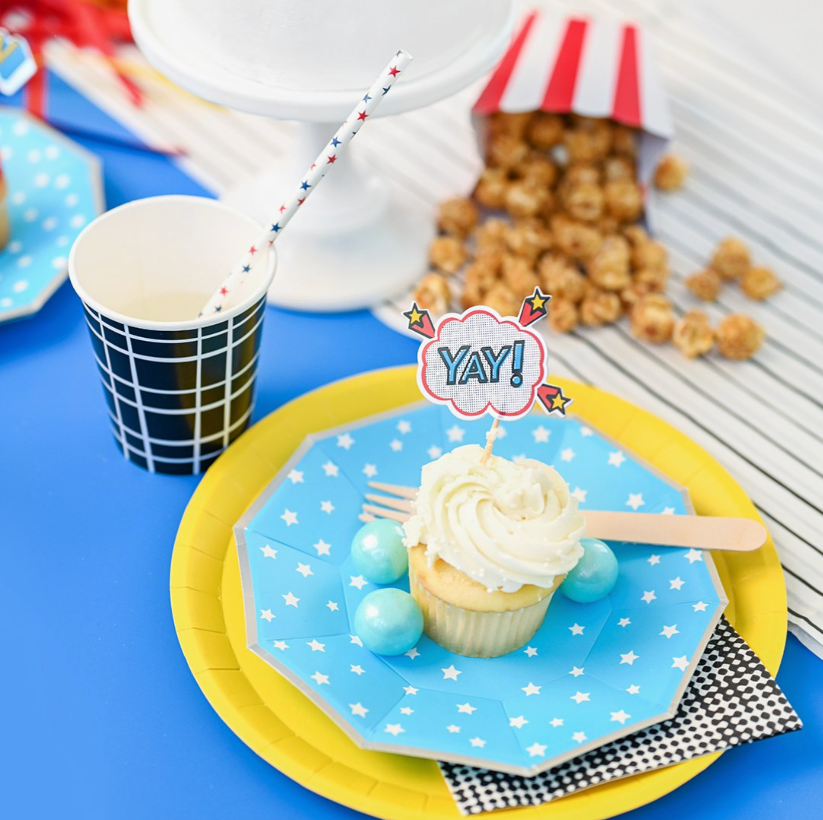 Cupcake on a blue plate with 'YAY!' topper, surrounded by party supplies on a blue tablecloth, including a paper straw with red and blue stars.