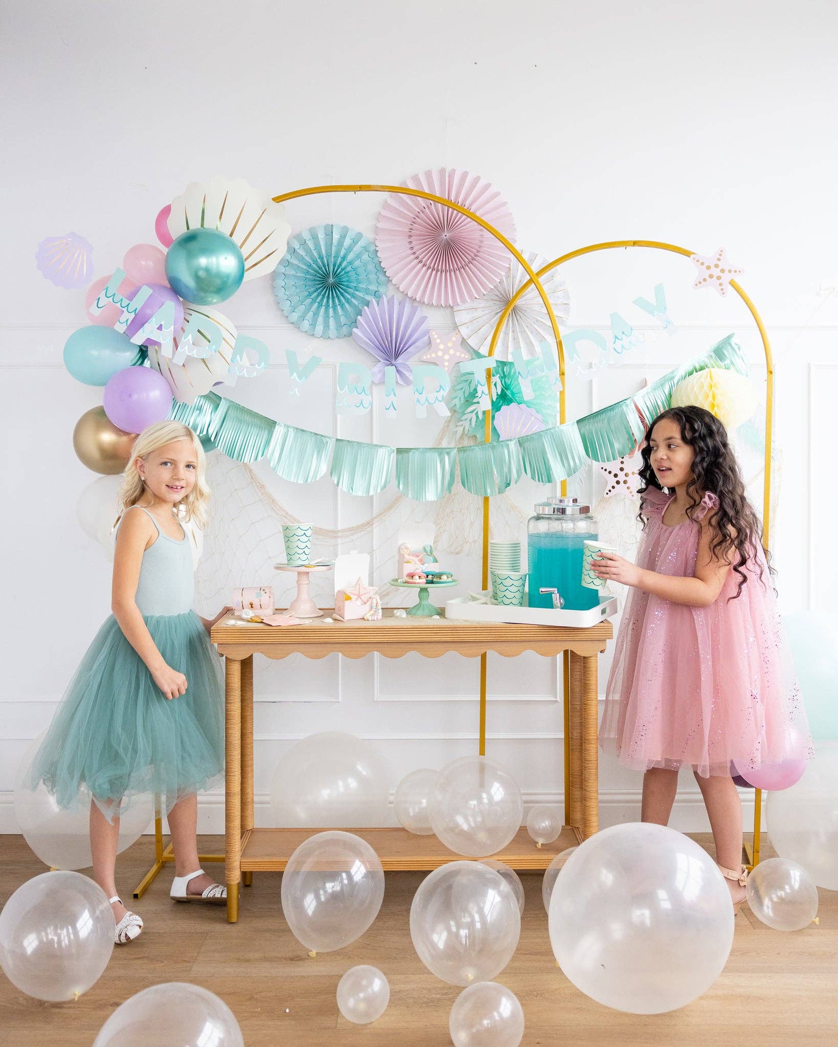 Two young girls in party attire standing near a mermaid theme decorated table with balloons and party decorations.