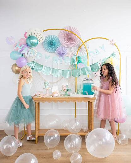 Two young girls in party attire standing near a mermaid theme decorated table with balloons and party decorations.