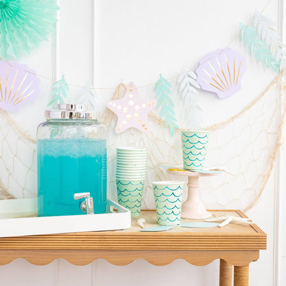 Mermaid-themed party setup with blue drink dispenser, green cups, and shell decorations.