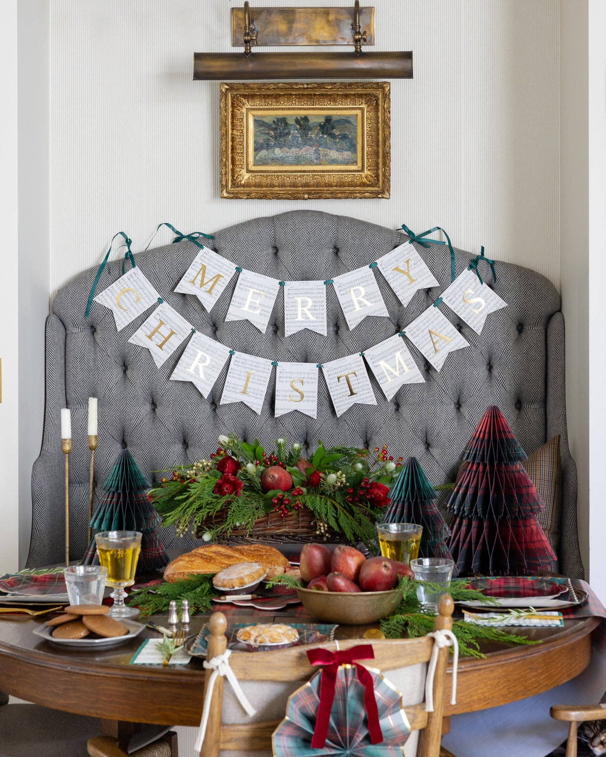 A festive holiday dining table setup in front of a gray tufted headboard, featuring a 'MERRY CHRISTMAS' banner made of hanging pennants, green garlands, red berries, small decorative trees, candles, and plated food.
