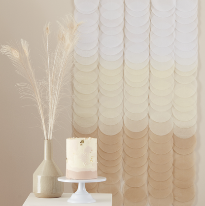 A neutral ombre tissue paper disc party backdrop displayed behind a cake and a vase of feathers, indicating use as a party decoration.