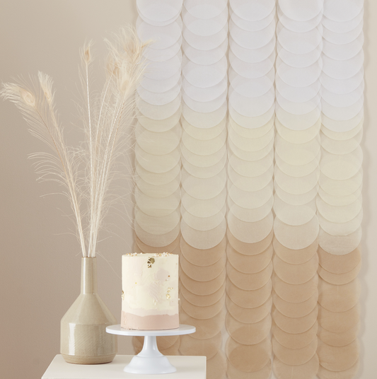 A neutral ombre tissue paper disc party backdrop displayed behind a cake and a vase of feathers, indicating use as a party decoration.