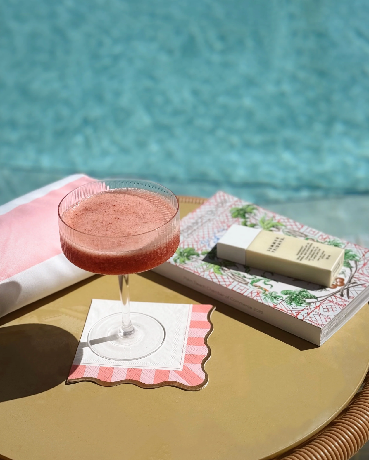 Cocktail glass with scalloped cocktail napkin on a table by a pool.