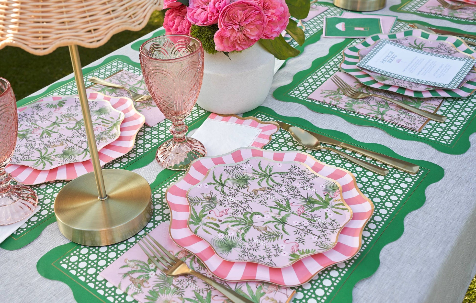 Decorative tropical table setting with pink and green plates, glasses, and flowers on patterned placemats.