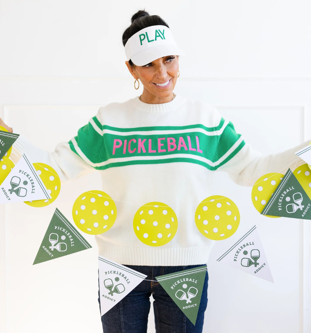 A person wearing a pickleball-themed sweatshirt with the word 'PLAY' on a headband, holding up two banners with pickleball designs.