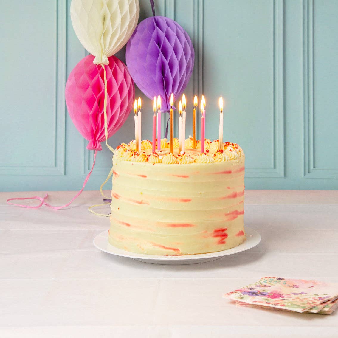 Birthday cake with lit candles on a table with colorful balloons against a light blue wall.