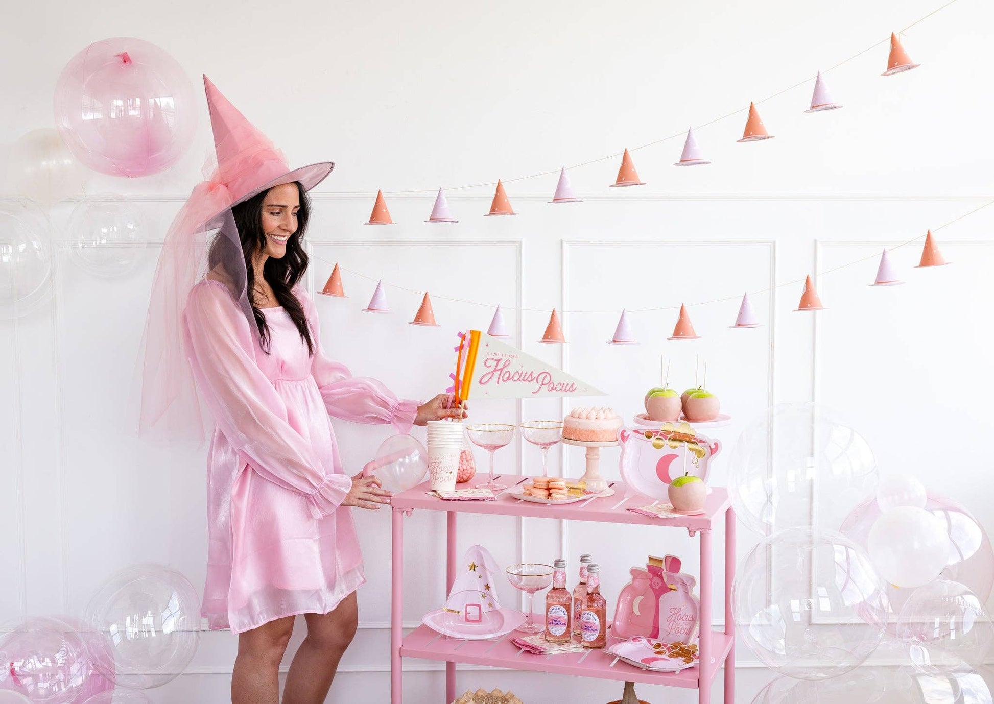 Woman in pink witch costume at a Halloween party standing next to a pink bar cart with drinks and snacks.