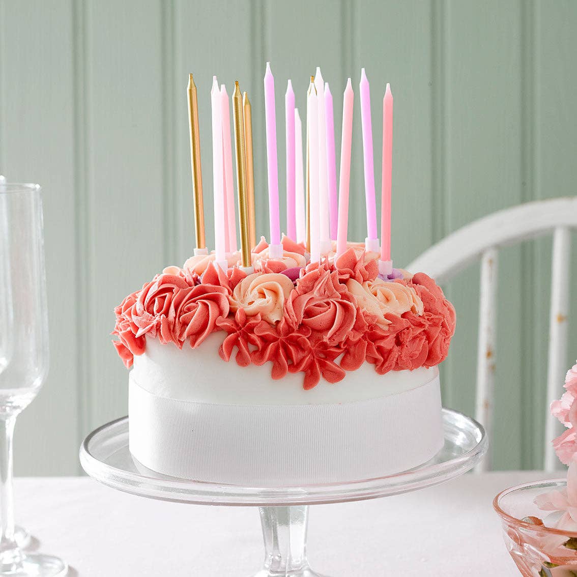 White cake with assorted pink and gold candles on a table with a light green wall background.