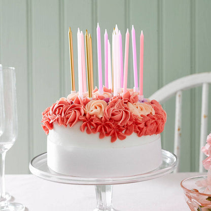 White cake with assorted pink and gold candles on a table with a light green wall background.