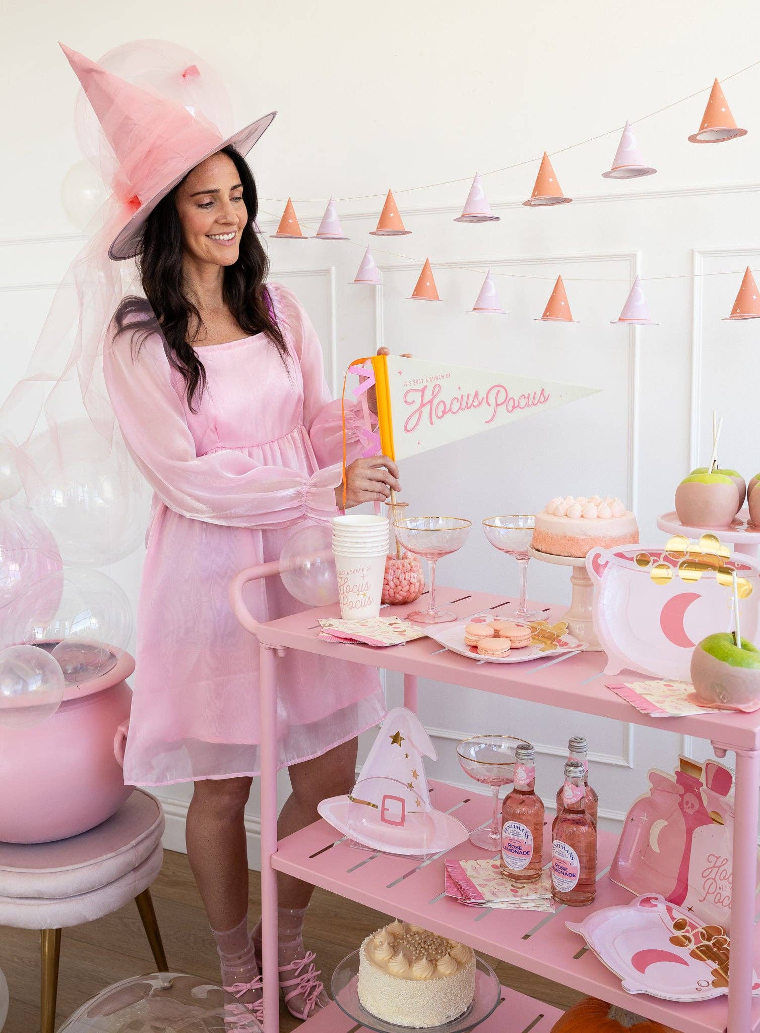 Woman in a pink witch costume and party hat at a pink-themed party with cake and drinks.