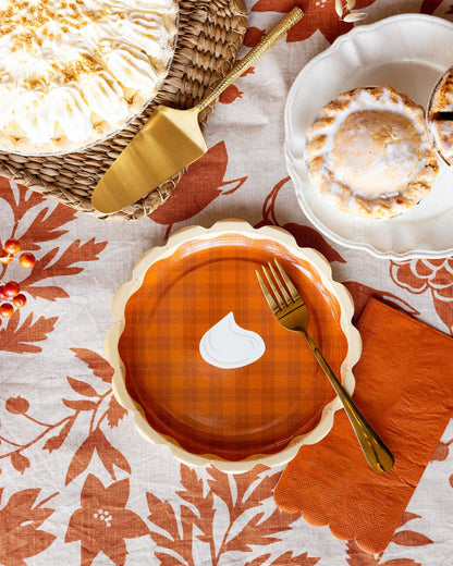 A decorative paper plate shaped like a plaid pumpkin pie on a with a gold fork on a patterned tablecloth.
