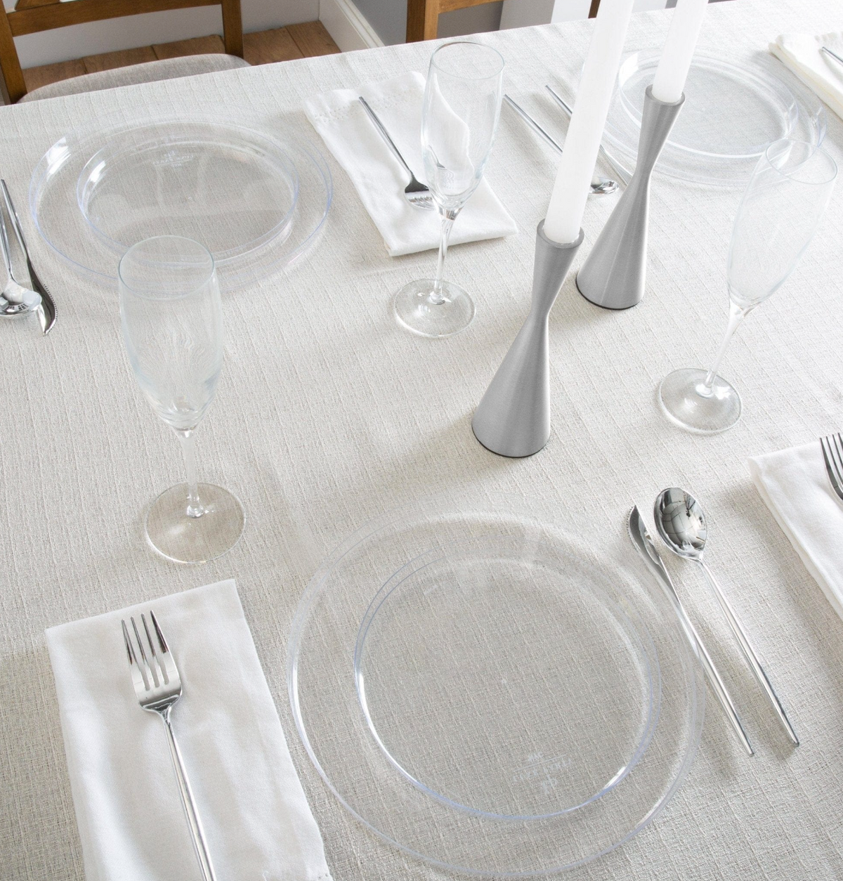 A styled dining table setting with clear plastic plates, white napkins, silvern plastic flatware, and silver candlesticks on a light beige textured tablecloth.