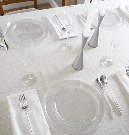 A styled dining table setting with clear plastic plates, white napkins, silvern plastic flatware, and silver candlesticks on a light beige textured tablecloth.