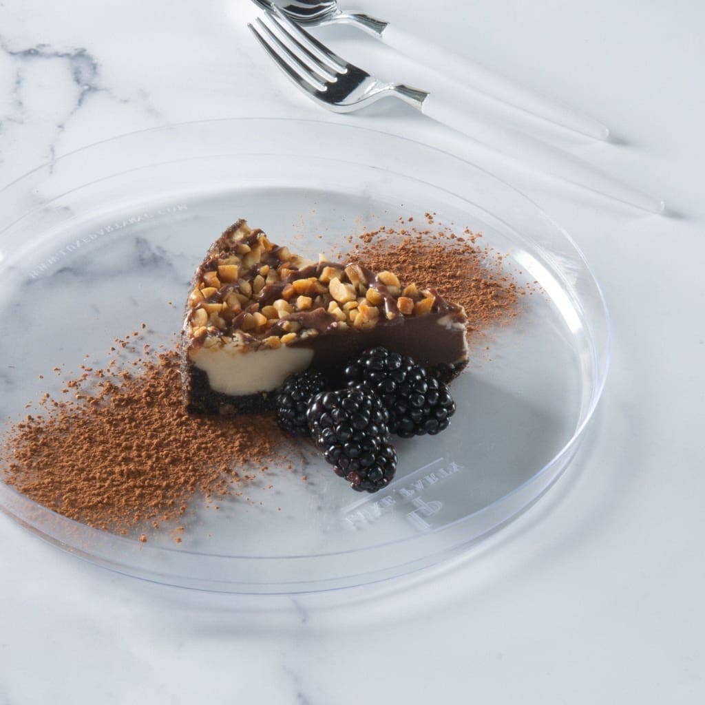 A clear round small plate on a white marble surface holding a slice of chocolate dessert topped with chopped nuts, with blackberries and cocoa powder garnish, and white-handled forks in the background.