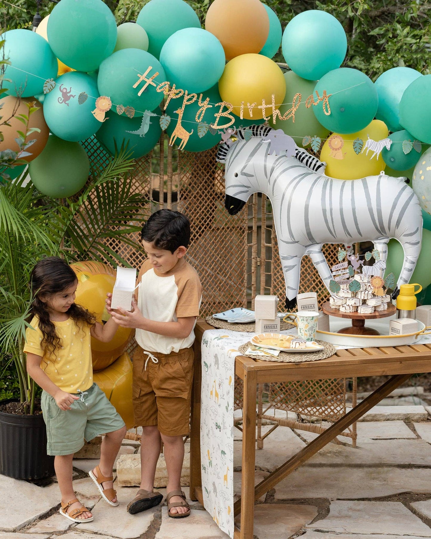 Children at a birthday party with balloons, a zebra decoration, and a table set with food.
