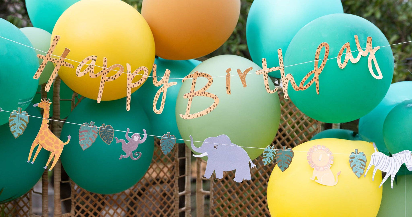 A birthday banner with the words 'Happy Birthday' and safari animal motifs, including a giraffe, placed against a blurred green and yellow background.