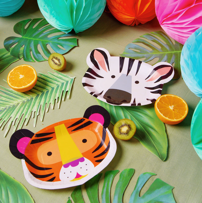 Tropical-themed paper party decorations including zebra and tiger face paper plates, green paper leaves, kiwi slices, orange wedges, and colorful paper lanterns arranged on a light green surface.