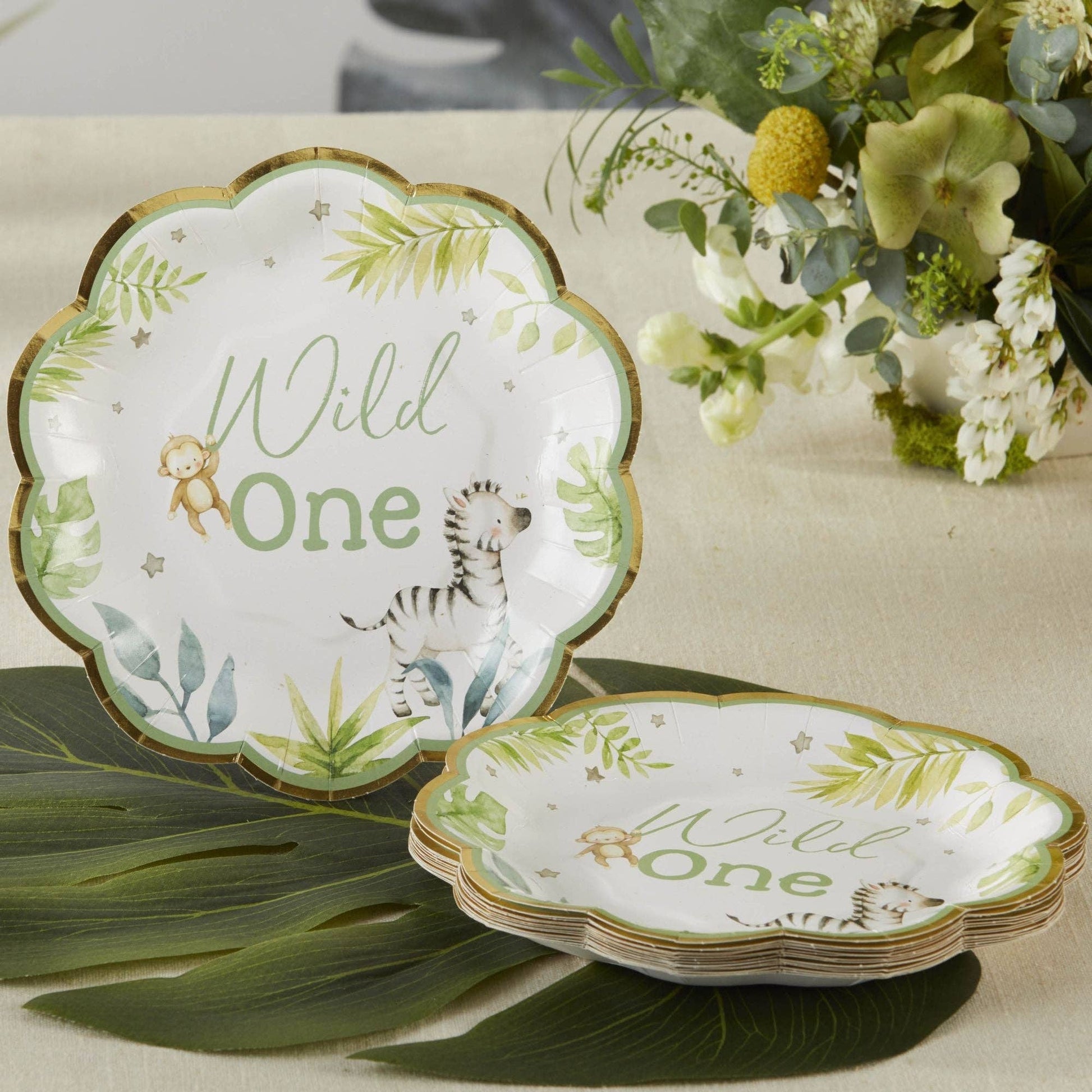 Set of 'Wild One' small paper plates with safari animal illustrations on a table with flowers and leaves.
