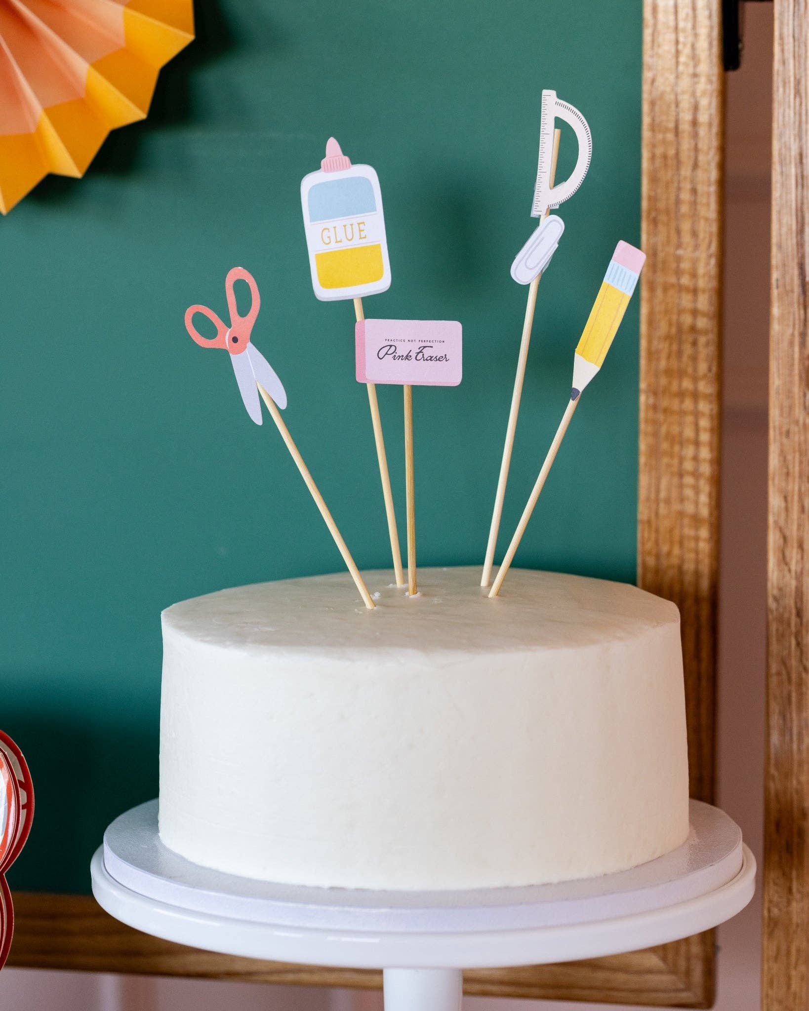 White cake with decorative school-themed toppers on a stand against a green chalkboard.