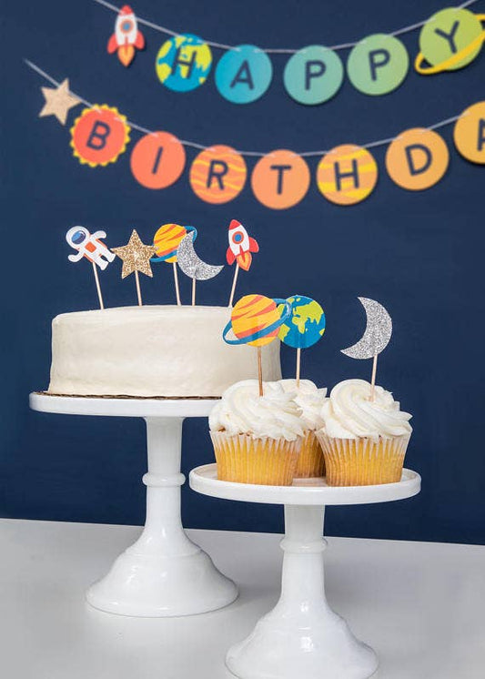 Cupcake toppers with astronaut, rocket, planet, and star designs, some glittered with gold and silver, displayed on cupcakes and a cake with a space-themed decoration in the background.