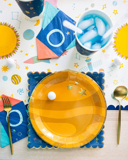 Colorful space-themed party table setting with yellow planet plates, blue cups, and gold utensils on a patterned tablecloth.