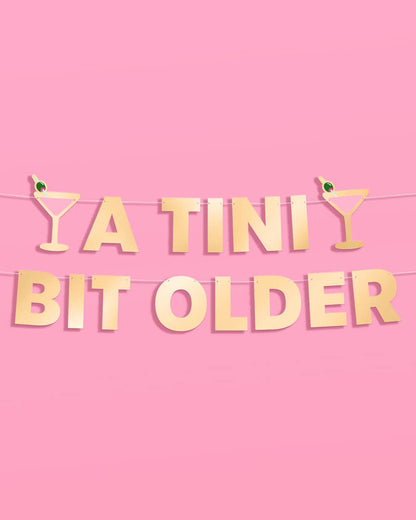Gold 'A TINI Bit Older' banner with cocktail glasses on a pink background