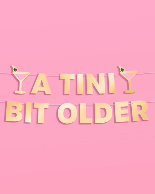 Gold 'A TINI Bit Older' banner with cocktail glasses on a pink background