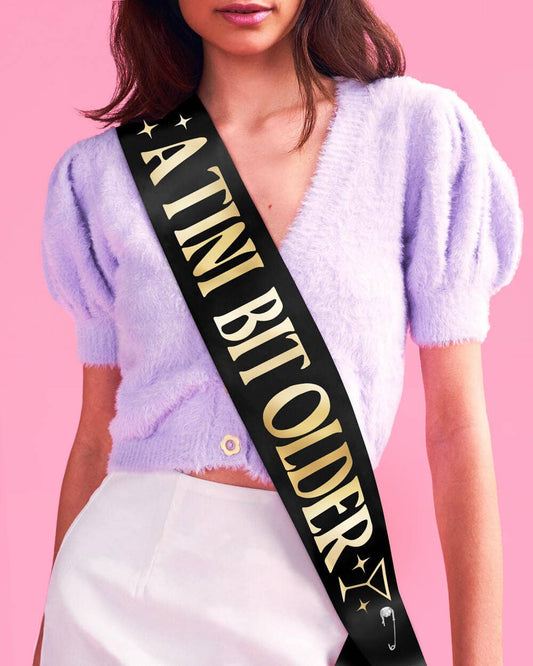 Person wearing a sash with 'A TINI BIT OLDER' text on a pink background