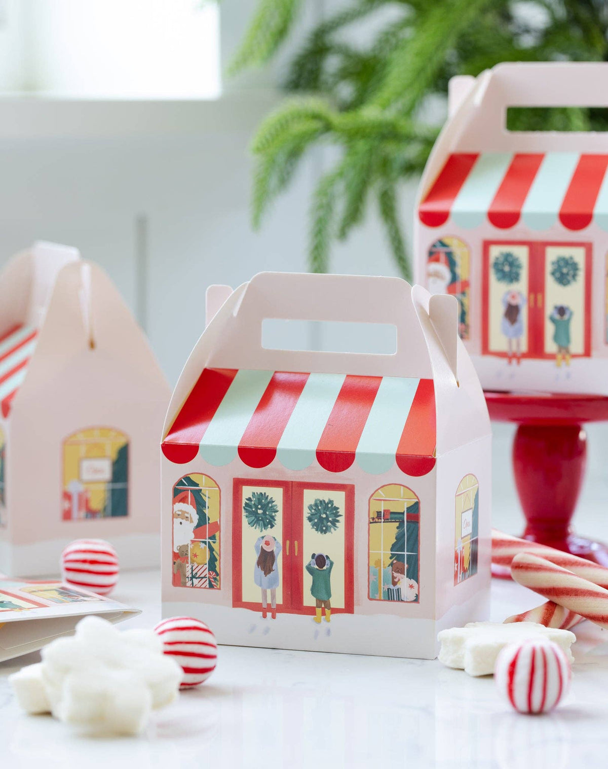 A set of light colored paper gift boxes decorated like a toy storefront, with red and mint green striped awnings and festive holiday illustrations, styled with candy on a bright white surface.