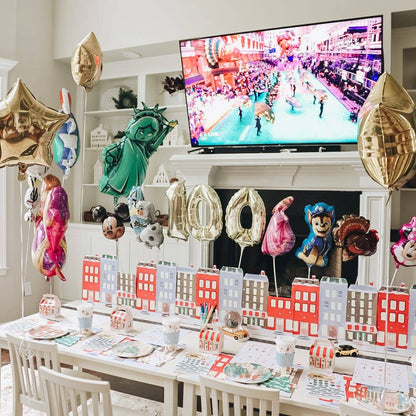 A decorated birthday party setup in a bright room, featuring a table with city-themed plates, cups, and paper buildings, large foil balloons including a Statue of Liberty balloon, and a TV mounted above a fireplace showing a parade scene.