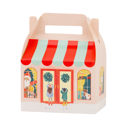 A light colored paper gift box shaped like a toy storefront, with a red and mint striped awning, illustrated shop windows, holiday wreaths, and a handle on top, shown on a white background.