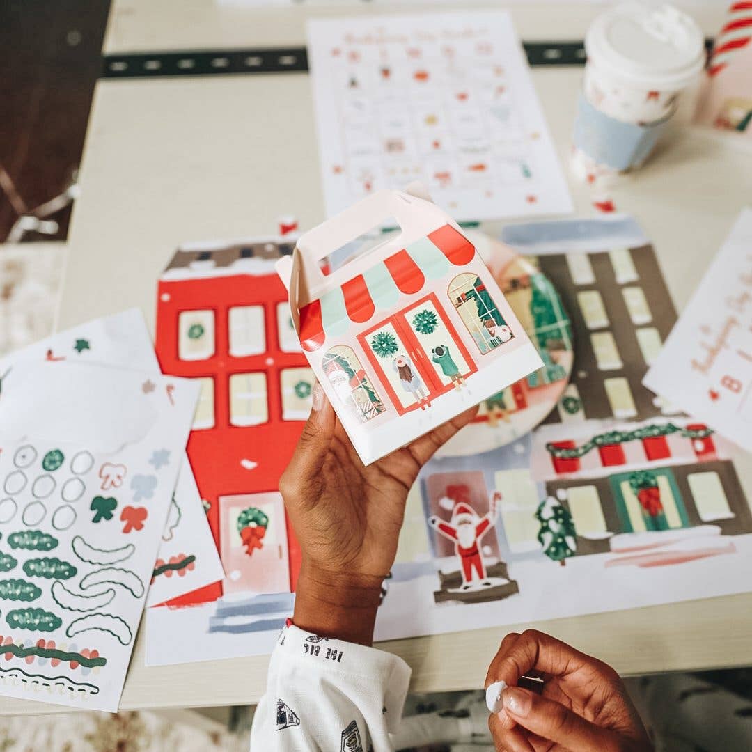 A person holds a small illustrated paper gift box with a holiday storefront design, surrounded by matching printed craft or sticker sheets on a table.