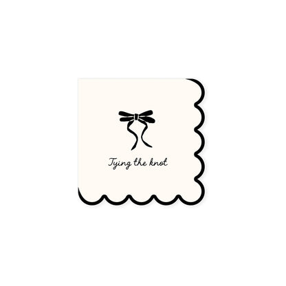 White scalloped cocktail napkin with a black bow design and text 'Tying the knot' on a white background.