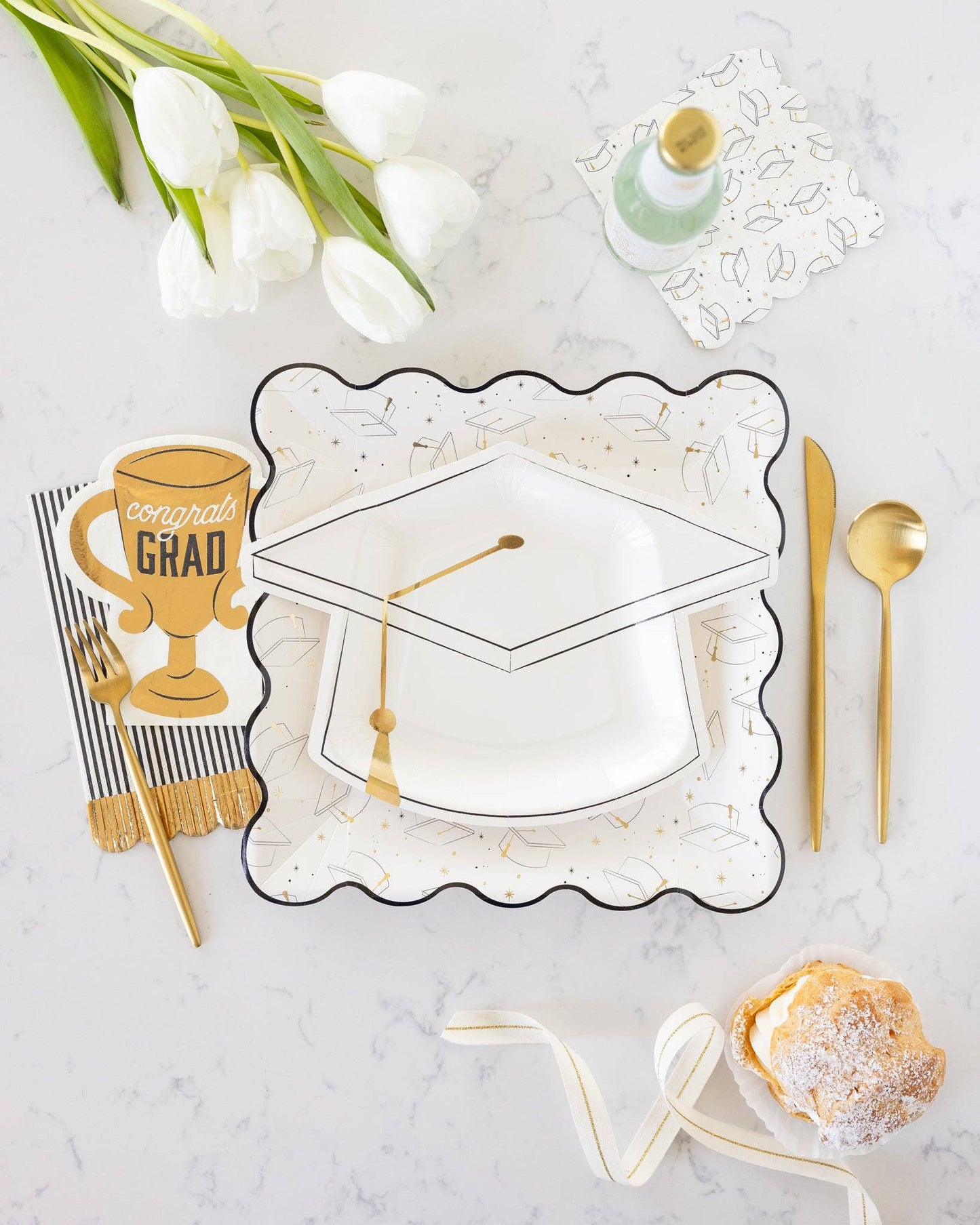 Graduation-themed table setting with white tulips, gold cutlery, and decorative plates on a marble surface.