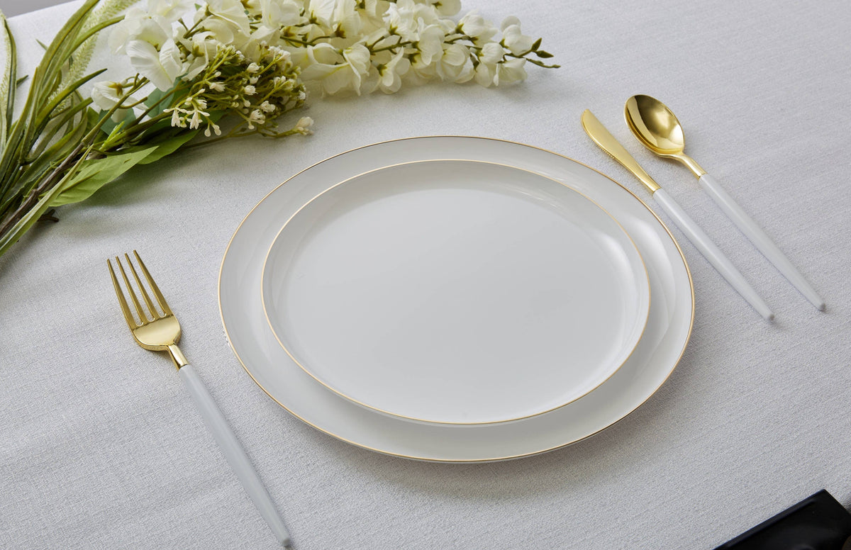 A place setting on a white tablecloth featuring a white plastic dinner plate with a delicate gold rim, accompanied by a gold-finished fork, knife, and spoon with white handles, styled with white flowers.